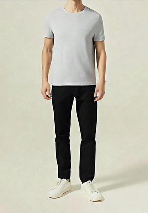 Homme portant un t-shirt gris clair uni à manches courtes, un pantalon noir et des baskets blanches, debout sur un fond clair.