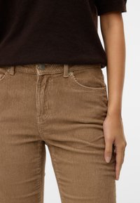 Gros plan d'une personne portant un pantalon en velours côtelé marron et un haut marron foncé, avec une main reposant à côté de la cuisse.