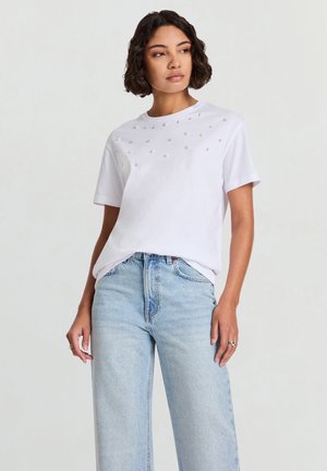 Maglietta bianca in cotone con maniche corte, decorata con borchie metalliche sulla parte anteriore. Abbinata a jeans a gamba larga in azzurro chiaro.