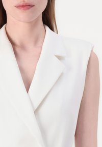 White sleeveless blazer with a notch lapel and a smooth, textured fabric. Features a deep V-neckline and structured shoulders.