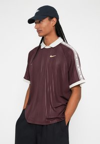 Polo shirt Nike em cor bordô com colarinho e detalhes nas mangas em branco. Apresenta um design de listras verticais e um pequeno logo dourado no peito.