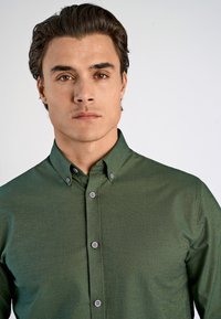 Camisa verde de botón fabricada con tela texturada, que presenta un cuello extendido y botones grises a lo largo del frente, exhibida en un modelo.