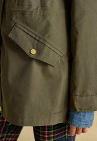 Zielona oliwkowa kurtka z boczną kieszenią ze złotymi elementami, gładką teksturą i widocznym logo. Pod spodem widoczny jest niebieski denimu rękaw.