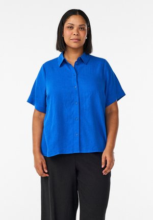 Kvinde med skulderlangt sort hår iført en lys blå kortærmet skjorte med knapper og sorte bukser, stående mod en hvid baggrund.