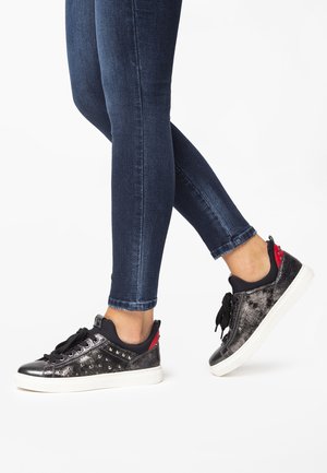 Zapatillas negras y plateadas con acentos metálicos y tachuelas; suela blanca; combinadas con jeans skinny azul oscuro. Presentan cordones negros y detalle en el talón rojo.