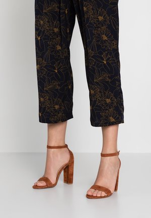 Brązowe zamszowe sandały na wysokim obcasie z paskami wokół kostki, z otwartym noskiem i grubym obcasem. W zestawieniu z ciemnymi spodniami w kwiatowy wzór 3/4.