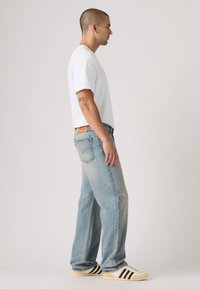 Λευκό βαμβακερό T-shirt, ανοιχτό μπλε τζιν με ξεθωριασμένο πλύσιμο και κρεμ αθλητικά παπούτσια με μαύρες ρίγες. Η πλάγια όψη δείχνει τη εφαρμογή και τις αναλογίες.