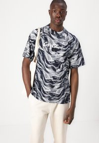 Short-sleeve navy t-shirt featuring a white wave pattern and "Café Colucci" text. The model holds a beige tote bag and wears light-colored pants.