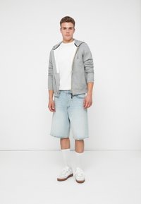 Młody mężczyzna stoi z neutralnym wyrazem twarzy, ubrany w szary rozpinany polar, biały T-shirt, jasne dżinsowe szorty, białe skarpetki i białe trampki.