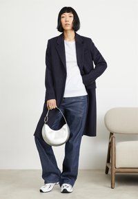 Marineblå lang frakke over en hvid t-shirt, parret med blå brede jeans, holder en sølv halvmåne håndtaske og bærer hvide sneakers.
