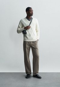 Pull en tricot blanc avec un motif en losanges, pantalon large en tweed marron et chaussures de ville noires. Petit sac à bandoulière noir.