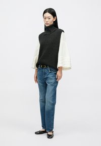 Grijze gebreide mouwloze trui met hoge kraag over een wit blouse met lange mouwen, gecombineerd met lichtblauwe spijkerbroek en zwarte platte schoenen.