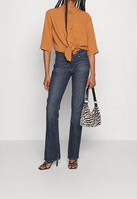 Camisa de color óxido, de mangas cortas y anudada en la cintura, combinada con jeans de mezclilla oscuros de pernera acampanada y un bolso de mano de estampado de cebra en blanco y negro.