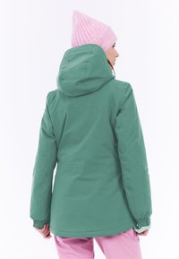 Chaqueta impermeable verde con capucha, puños elásticos y textura suave. Combinada con pantalones rosas, mostrando un look casual al aire libre.