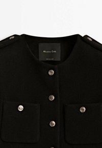 Veste en laine noire avec encolure ronde, boutons décoratifs argentés sur le devant et sur deux poches poitrine, et épaulettes aux épaules.