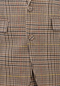 Kockás bézs, barna és fekete dzseki. Két zsebbel rendelkezik, és kiemelkedő sárga és piros varrással díszített, gomb részletekkel.