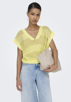 Vrouw draagt een losse gele gebreide top, lichtblauwe hoge taille jeans, grote oorringen en houdt een beige oversized handtas vast.