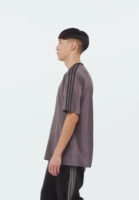 Šedé sportovní tričko s krátkým rukávem, s třemi černými pruhy na ramenou. Hladká textura a uvolněný střih.