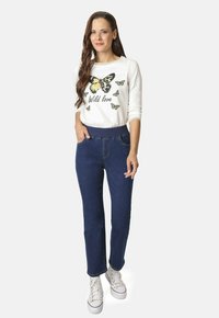 Camisa blanca de manga larga con gráficos de mariposas y texto "Amor salvaje", combinada con pantalones de mezclilla azul oscuro y zapatillas blancas.