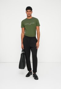 Grønn bomull T-skjorte med logo, svarte elastiske bukser, svarte lær-sko, holder en svart tote bag, står på en hvit bakgrunn.