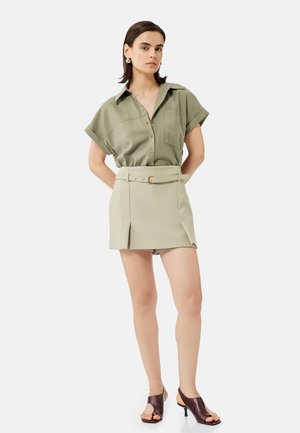 Nainen, jolla on lyhyet tummat hiukset ja yllään oliivinvihreä lyhythihainen napillinen paita, beige vyöllinen minihame ja ruskeat avokkaat, seisoo.