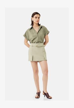 Mujer con pelo corto y oscuro que lleva una camisa de manga corta color verde oliva abotonada, minifalda beige con cinturón y tacones marrones de punta abierta, de pie.