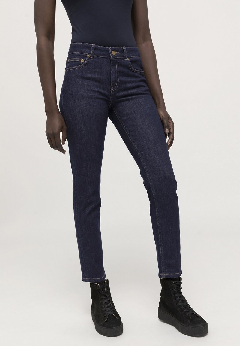 Dunkelblaue Denim-Skinny-Jeans mit hoher Taille, die mit goldenen Hardware-Akzenten und einer glatten Textur versehen sind. Getragen mit schwarzen Schnürstiefeln.