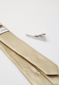 Cravate en soie beige avec une texture lisse, accompagnée d'un pince cravate en métal argenté présentant un design de vague.