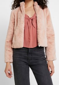 Nepbont jas in zachtroze, met een ritssluiting, hoge kraag en geribbelde manchetten. Gedragen over een gestructureerde, blush top met een strik aan de voorkant.