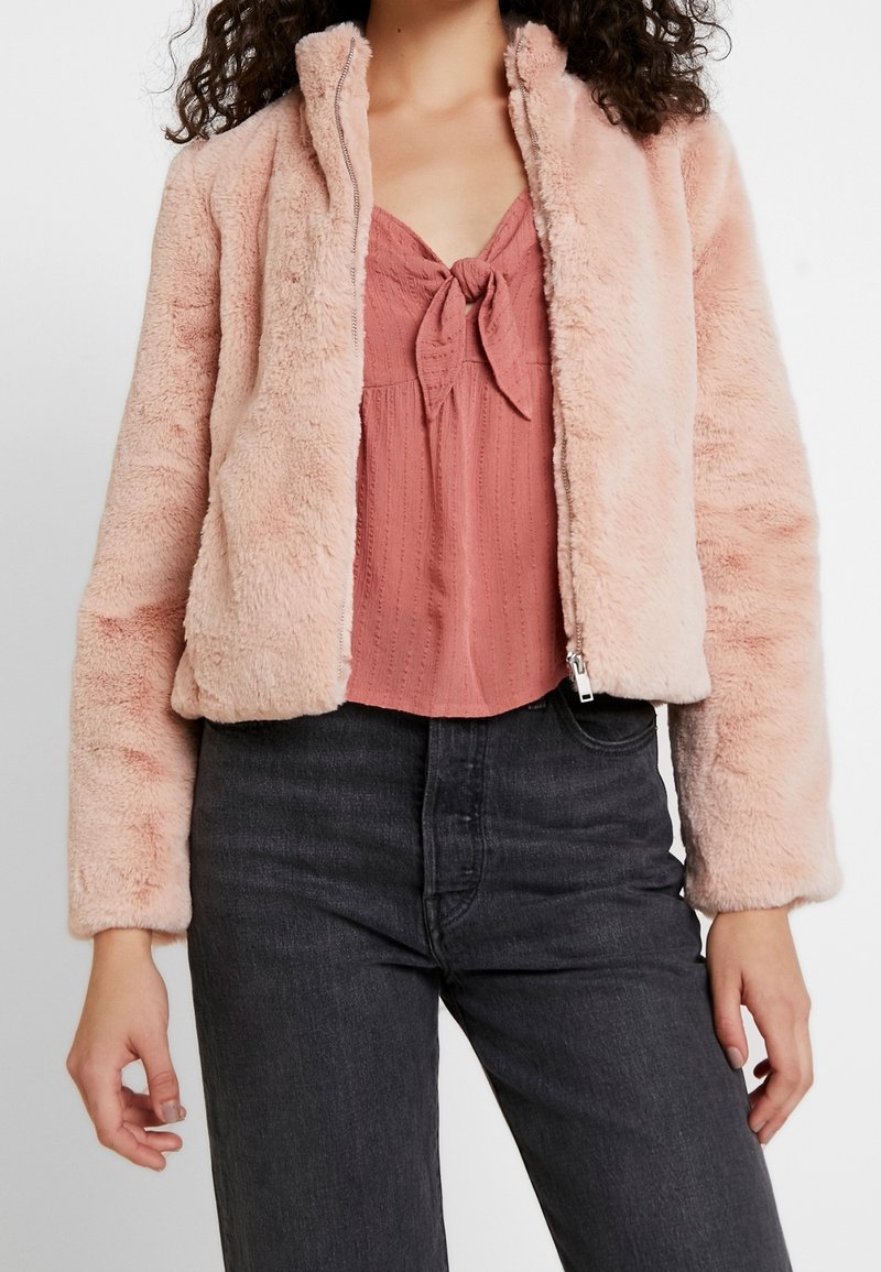 Nepbont jas in zachtroze, met een ritssluiting, hoge kraag en geribbelde manchetten. Gedragen over een gestructureerde, blush top met een strik aan de voorkant.