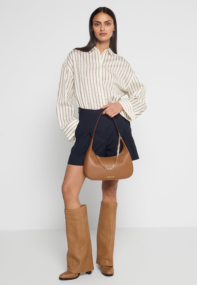 Camicia oversize a righe beige e marrone, pantaloni corti blu navy, stivali alti marroni e una borsa marrone con tracolla dorata. Tessuto liscio e vestibilità comoda.