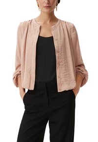 Jasnoróżowa, lekka bluzka z okrągłym dekoltem, zapinana na guziki, z bufkami na rękawach, noszona na czarnym topie i zestawiona z czarnymi spodniami.