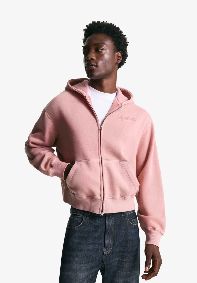 Lyserød zip-up hættetrøje i blødt stof, med en frontlomme, justerbar hætte og ribkant. Bæres over en hvid t-shirt.