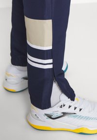 Navy athletic pants with beige accents and white stripes, featuring zipper openings at the ankles. Paired with white and yellow sports shoes.
