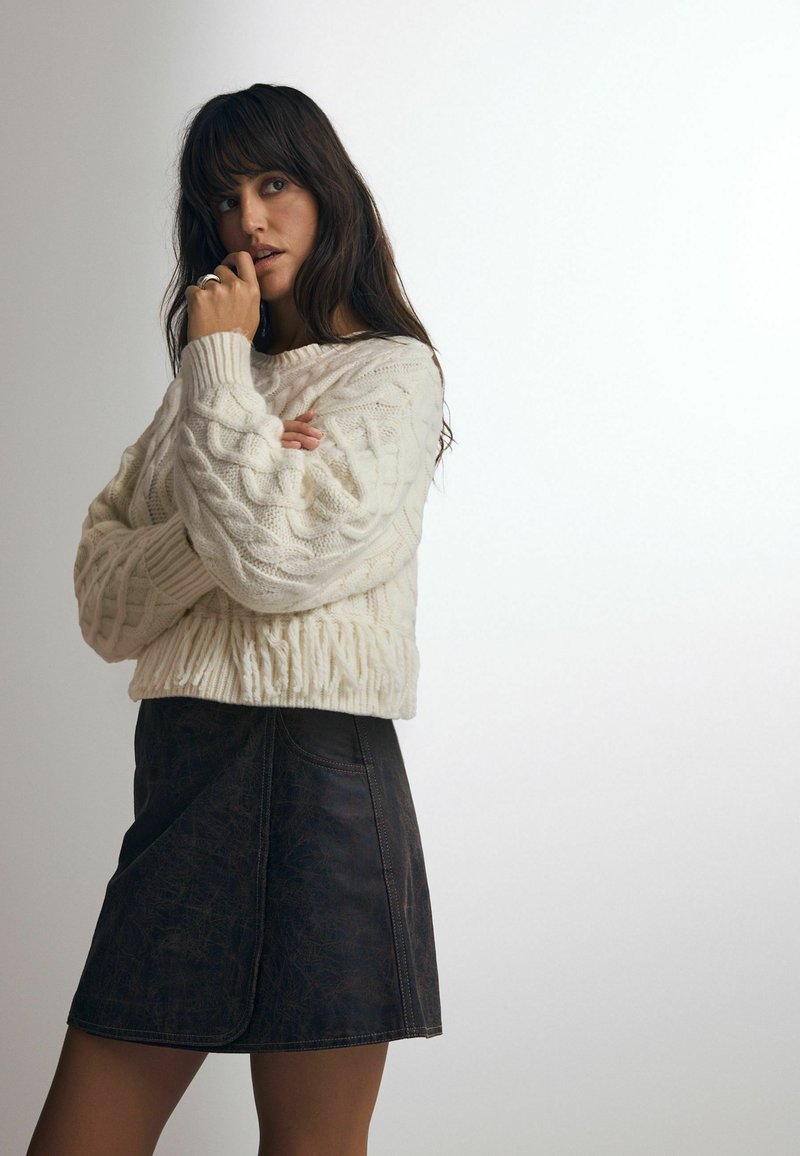 Cremfarbener Kabelstrickpullover mit Rippensaum, kombiniert mit einem dunkelbraunen Kunstlederminirock mit einer diagonalen Vordernaht.