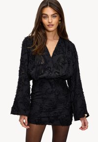 Zwarte jurk met lange mouwen, textuurstof met verheven patronen, diepe V-hals, aansluitende taille en uitlopende mouwhems.