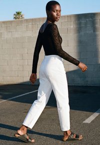 Jonge vrouw die buiten loopt, gekleed in een zwarte lange mouw top met luipaardprint, witte hoge taille broek en zwarte sandalen.