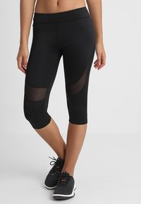 Nainen, jolla on yllään mustat lyhyet legginssit, joissa on verkkopaneeleja reisissä ja mustat urheilukengät, seisoo yksinkertaista taustaa vasten.