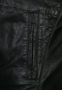 Pelle nera con una texture liscia, caratterizzata da una cucitura verticale e un dettaglio con zip nascosta. La superficie presenta leggere pieghe e una grana naturale.