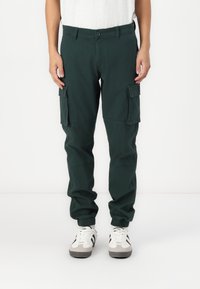 Cargo-Hosen aus dunkelgrünem Baumwollmaterial mit elastischen Bündchen, die mehrere Seitentaschen, einen normalen Schnitt und einen Knopfverschluss aufweisen. Kombiniert mit Turnschuhen.