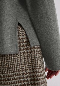 Grijze gevoerde top met openingen aan de zoom, gelaagd over een stof met een bruine en beige houndstooth patroon. Zachte textuur zichtbaar in de materialen.