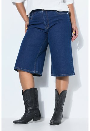 Person iført mørkeblå denimshorts til knæene, sorte cowboystøvler, hvid top, med hænderne langs siderne og gul neglelak.