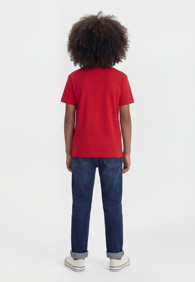 Niño con cabello rizado que lleva una camiseta roja de manga corta, vaqueros azules remangados y zapatillas blancas, de pie mirando hacia un fondo blanco liso.