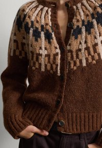 Bruine gebreide cardigan met een ronde hals, knoopsluiting, ingewikkeld patroon bij de schouders in beige en donkerblauw, en een geribbelde onderrand.