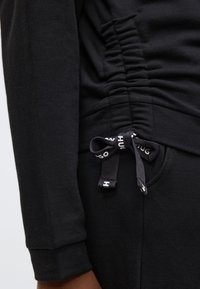 Musta collegepaita, jossa on kerättyjä yksityiskohtia sivuissa ja kiristysnauhat. Pehmeä tekstuuri ja kontrastivärinen valkoinen logo nauhoissa.