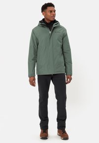 Giacca impermeabile verde con cappuccio, chiusura con zip e polsini elasticizzati. Abbinata a pantaloni neri e scarponi da trekking marroni.