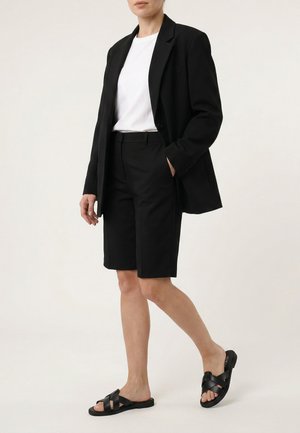 Kvinde iført en sort blazer, sorte knælange shorts, hvid t-shirt og sorte flade sandaler, stående mod en ensfarvet baggrund.