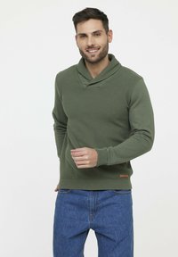 Grüner Strickpullover mit Kragen, gerippten Bündchen und Saum. Strukturierter Körper und brauner Lederakzent an der unteren linken Seite.