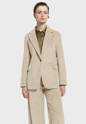 Femme portant un costume en velours côtelé beige avec un blazer à un bouton et un pantalon large assorti, sur une chemise boutonnée vert olive.