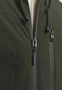 Chaqueta de color verde oscuro que muestra un primer plano de un cierre frontal con cremallera y herrajes negros, con cordones ajustables, fabricada en un tejido suave y mate.